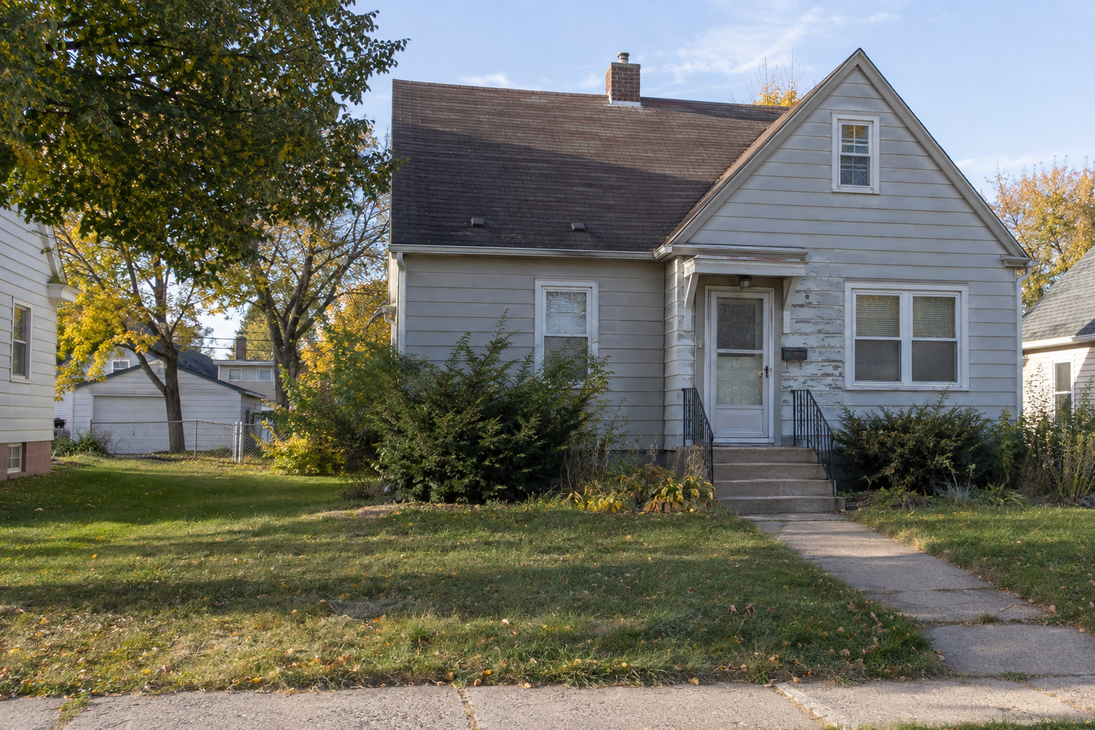 A modest older house with weathered siding, overgrown shrubs, and a cracked walkway.